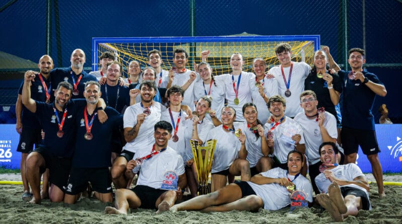 A beach soccer team poses triumphantly on the sand with their medals and trophy, celebrating a victory under stadium lights.