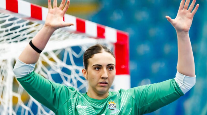 A goalkeeper in a green jersey raises both hands in a defensive stance in front of a red-and-white goal.