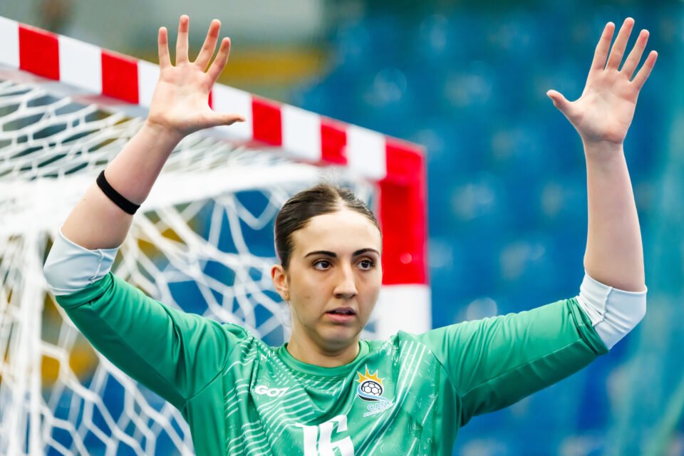 A goalkeeper in a green jersey raises both hands in a defensive stance in front of a red-and-white goal.
