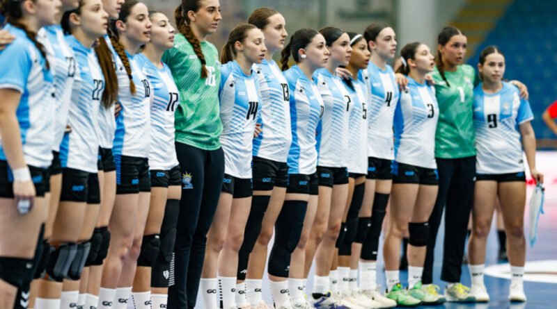 A group of athletes in matching uniforms stand in a line on an indoor court, with one player in a green jersey positioned slightly apart.